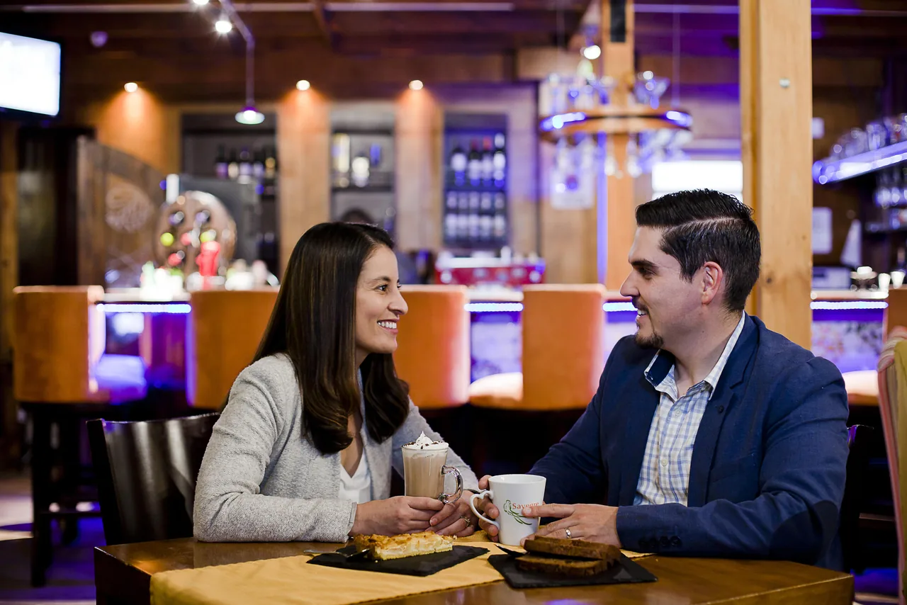 Una pareja conversa en una mesa del bar, con dos cafés y un postre delante y la barra iluminada al fondo