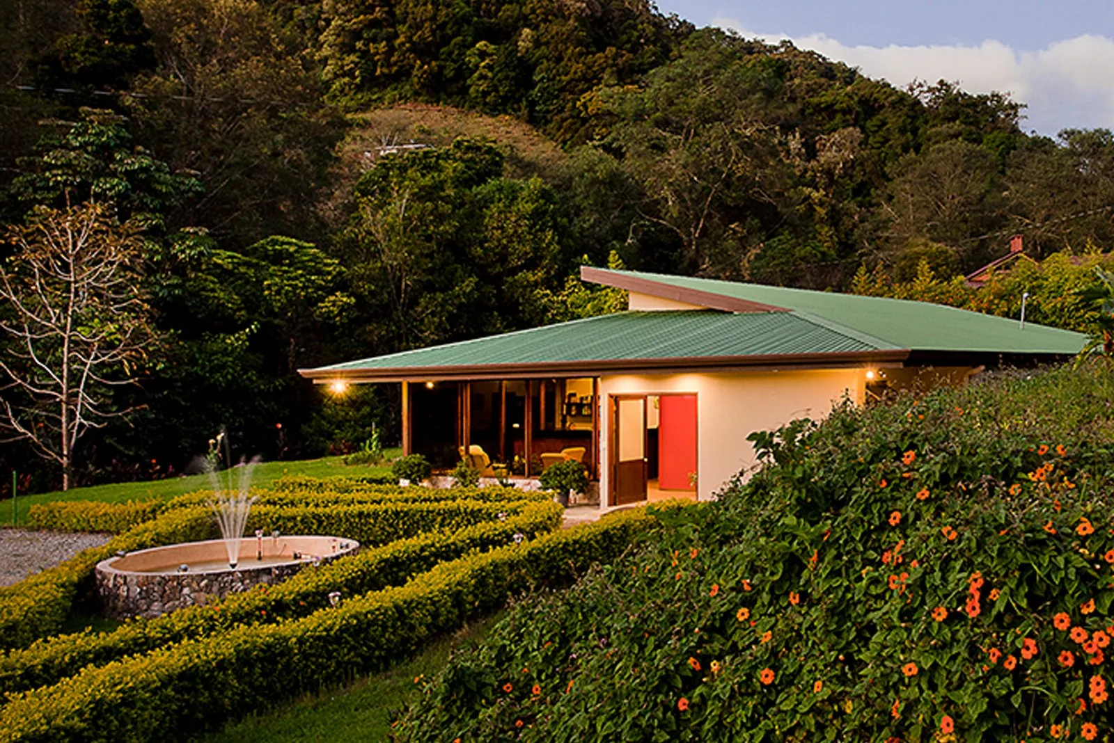 El edificio del Del Río Spa al atardecer, con los setos recortados en curva, una fuente redonda y macizos de flores naranjas delante