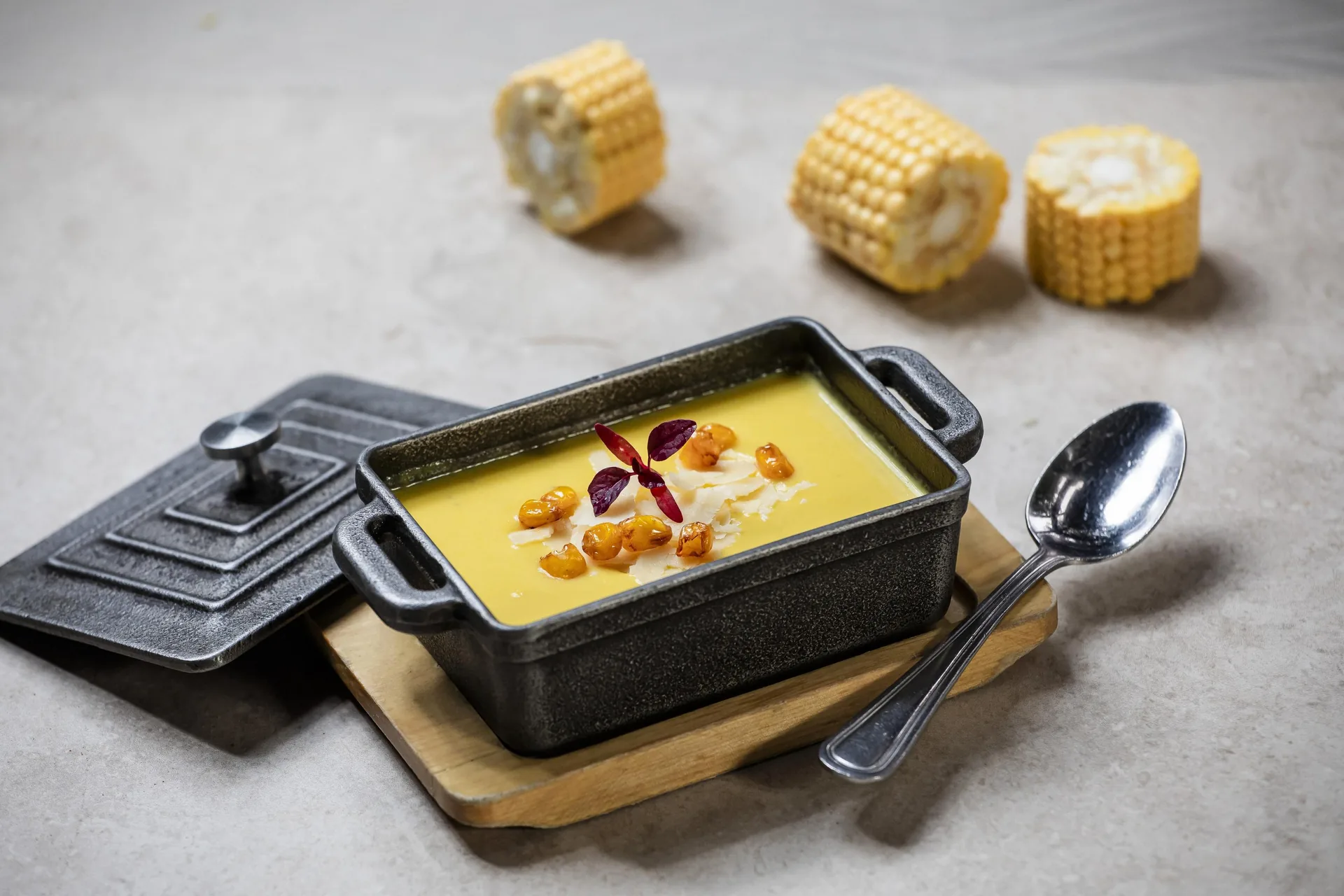 Crema amarilla en una fuente rectangular de hierro, con granos tostados, hebras de queso y un pétalo rojo por encima; al lado, la tapa, una cuchara y tres trozos de mazorca.
