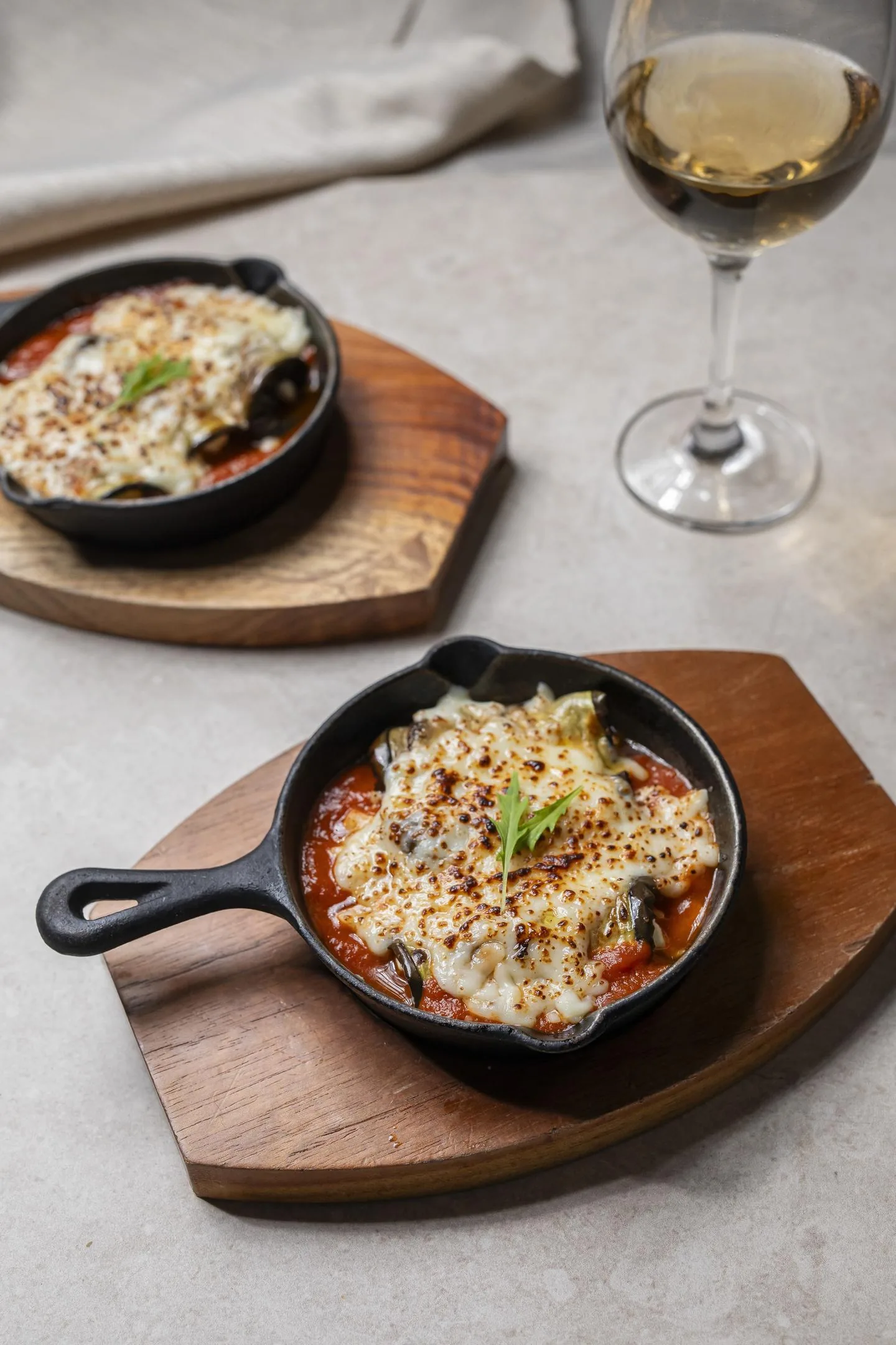 Gratinado de queso fundido sobre salsa de tomate en dos sartenes pequeñas de hierro, con una copa de vino blanco al lado.