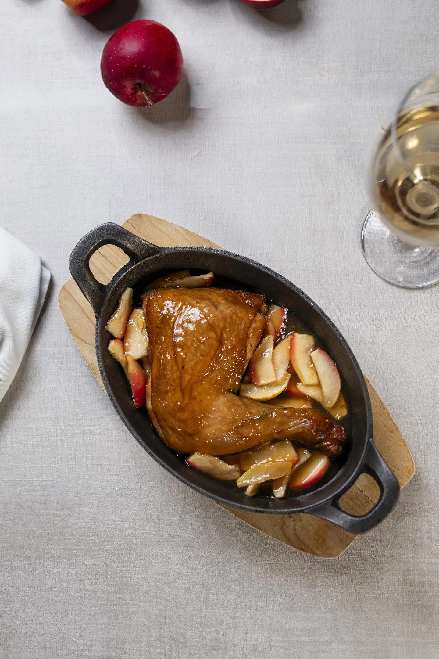 Cuarto de pollo de piel dorada rodeado de gajos de manzana cocidos, en una fuente ovalada de hierro; al lado, manzanas rojas y una copa de vino blanco.