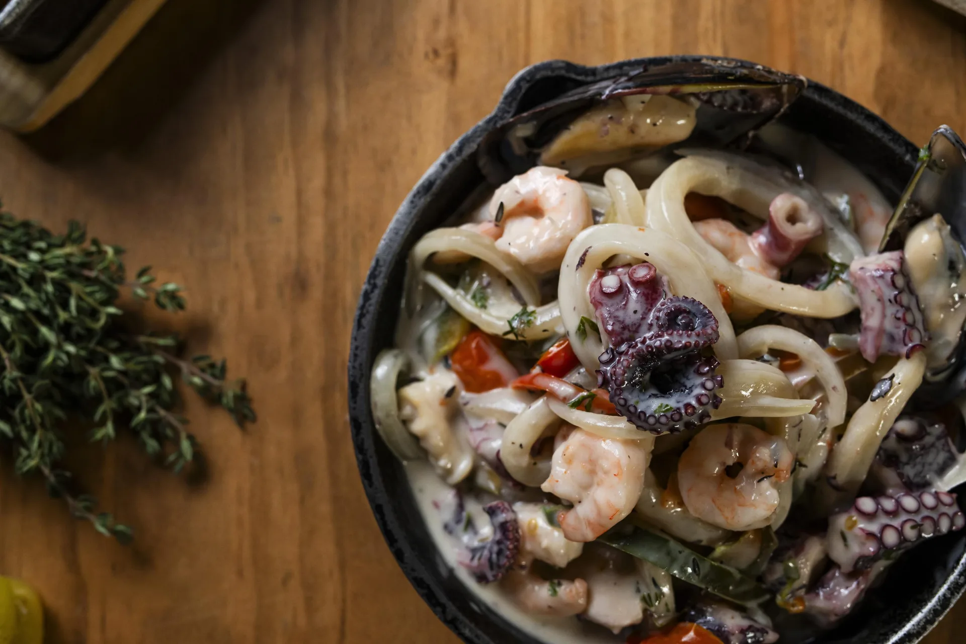 Guiso cremoso de mariscos en un bol negro, con camarones, aros de calamar, pulpo y un mejillón en su concha; a la izquierda, un manojo de tomillo sobre la mesa de madera.