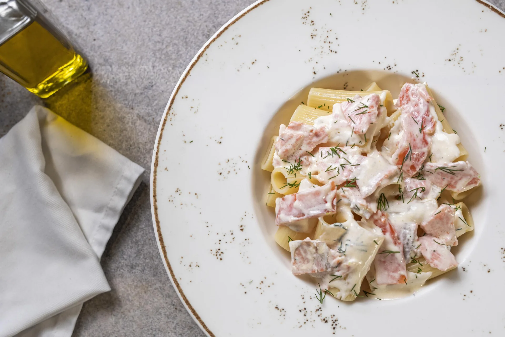 Pasta corta en salsa blanca con trozos de pescado rosado y eneldo, en un plato hondo de borde dorado, junto a una aceitera de vidrio.