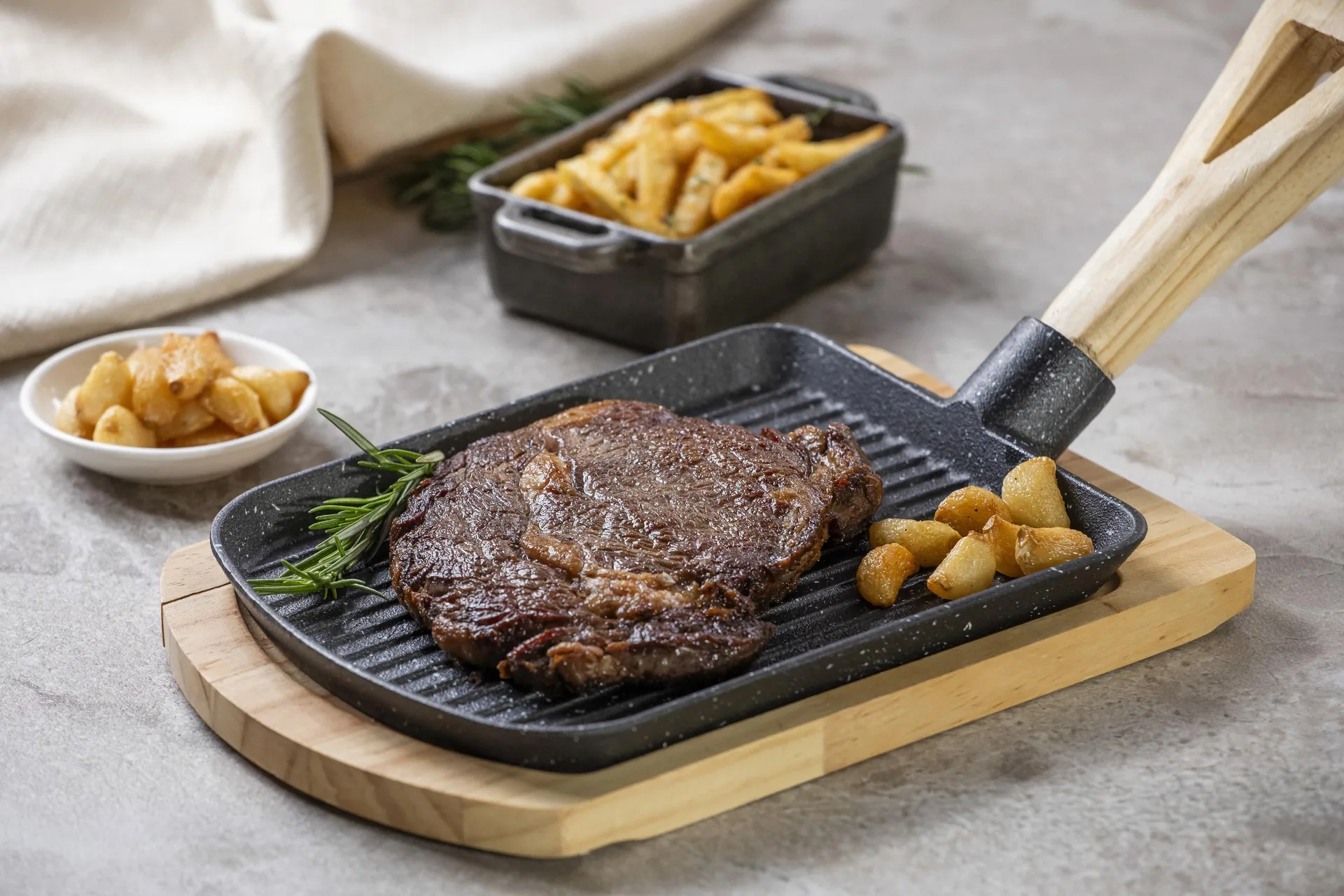 Corte de res a la parrilla con marcas de asado, servido en una plancha de hierro con romero y papas doradas; al fondo, dos fuentes más de papas.