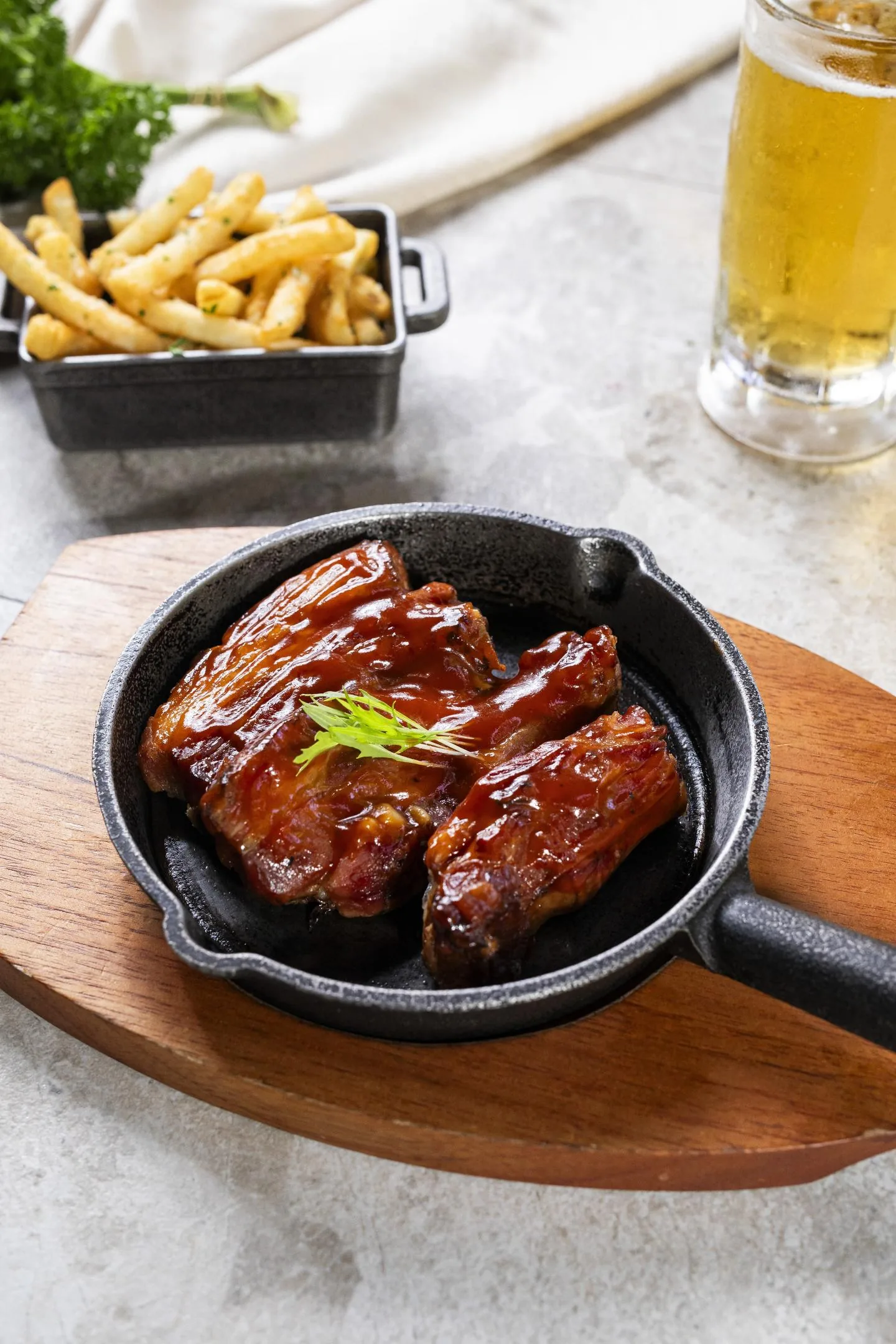 Costillas de cerdo glaseadas en una sartén de hierro sobre tabla de madera, vistas desde arriba, con papas fritas y una cerveza al fondo