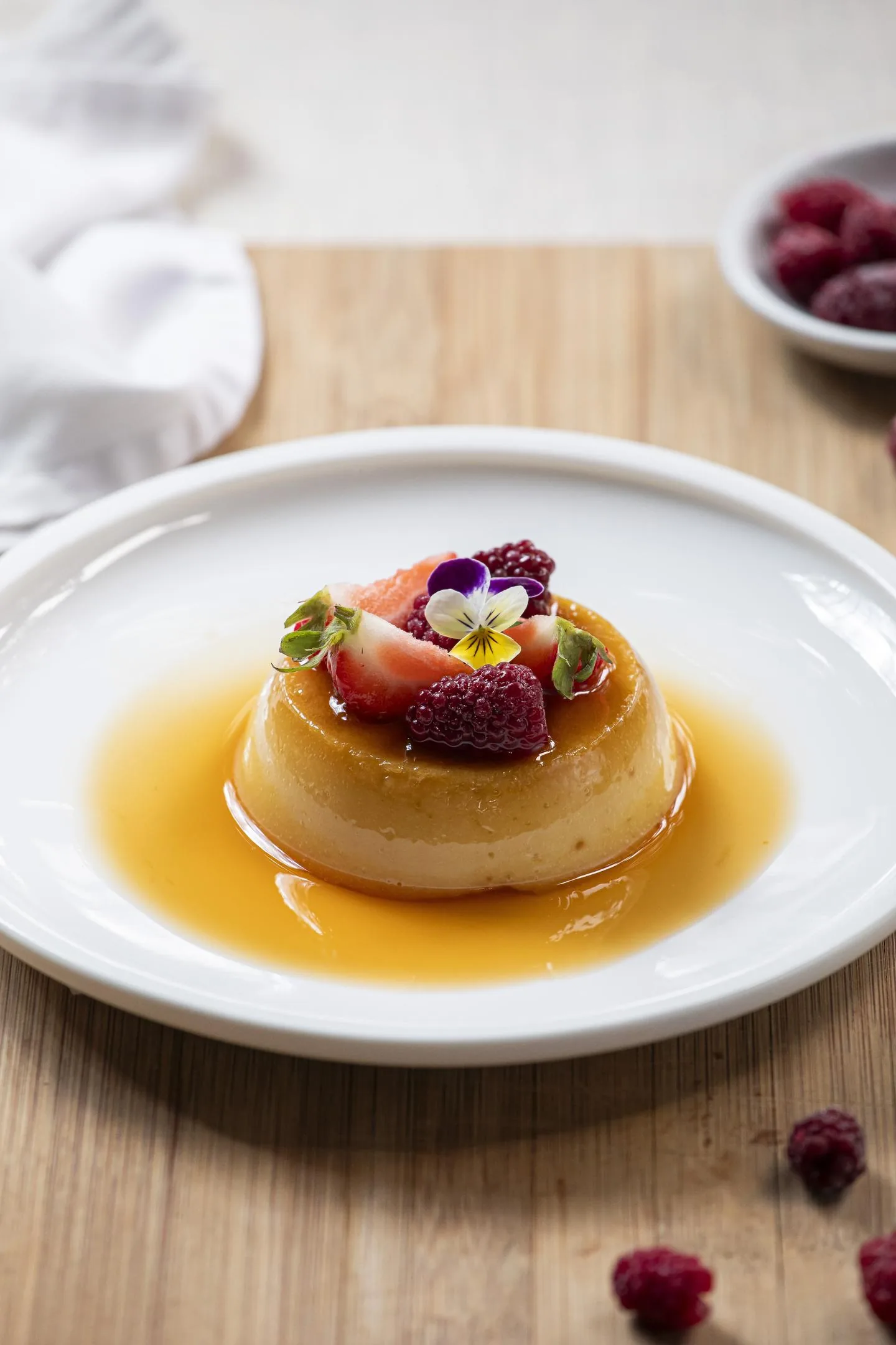 Flan bañado en caramelo, coronado con frambuesas, una flor comestible y hojas verdes; al lado, un cuenco de frambuesas.