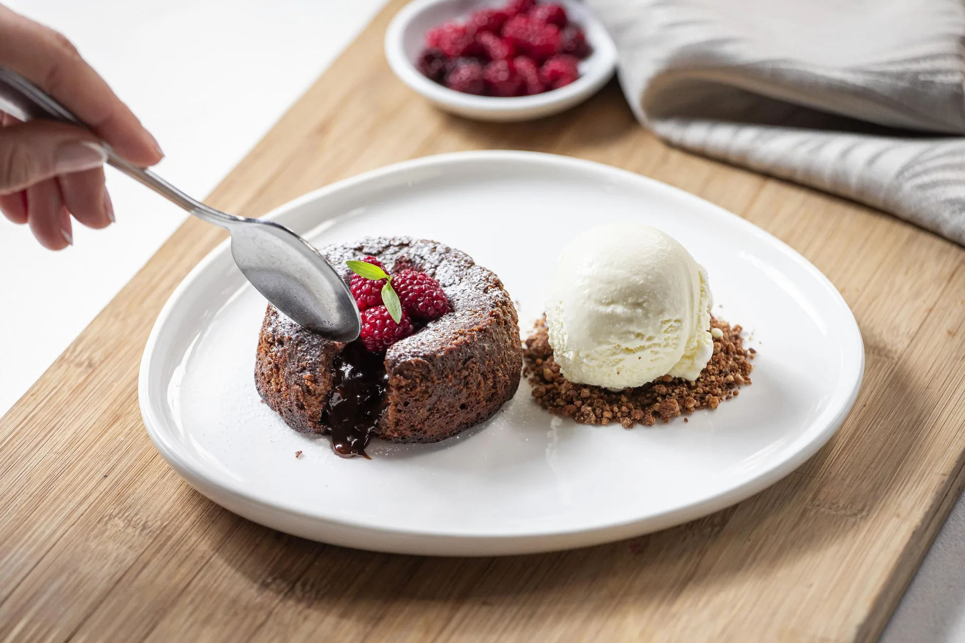 Una mano abre con la cuchara un volcán de chocolate y el relleno se derrama; al lado, una bola de helado sobre migas de galleta, con frambuesas y menta.