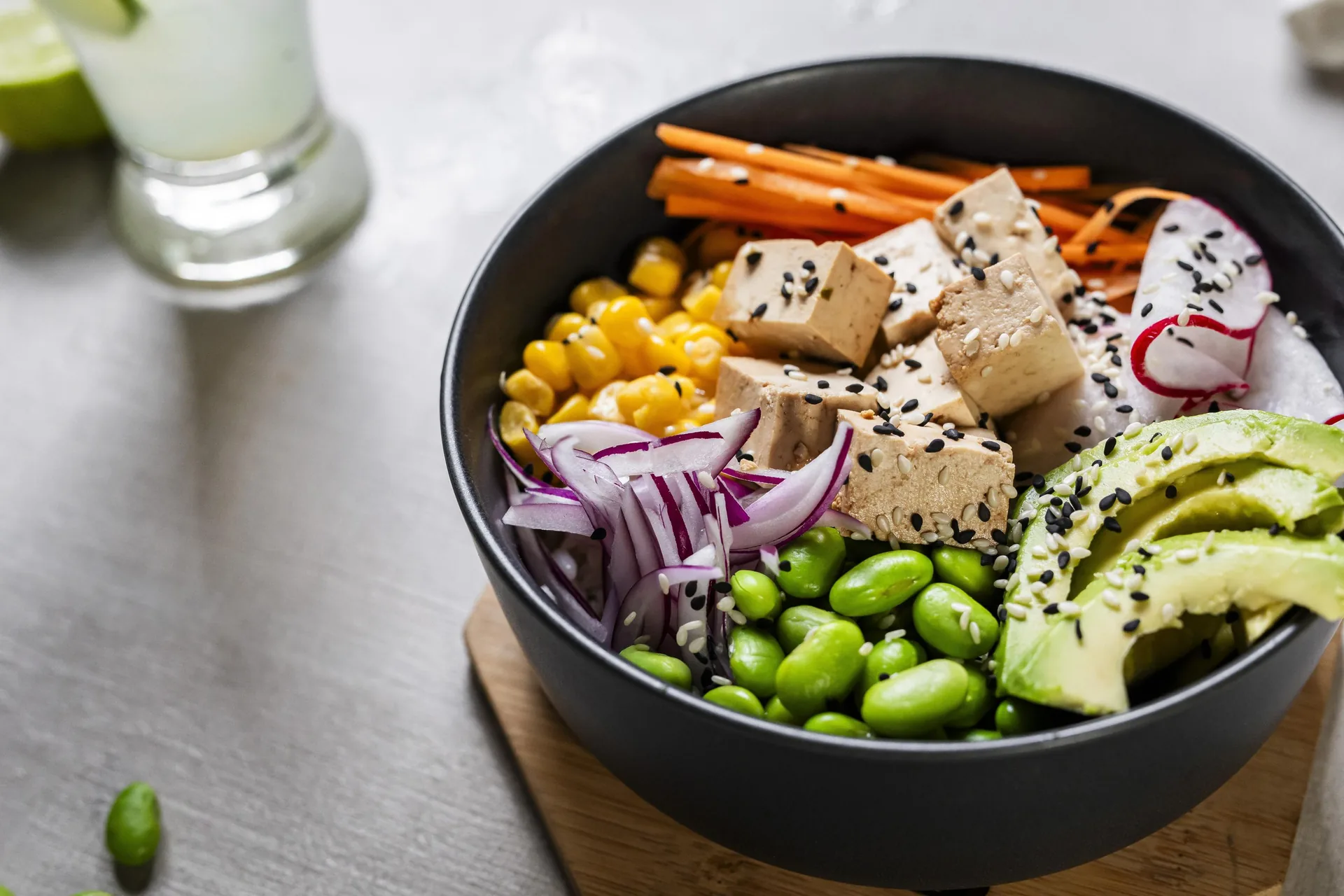 Bol de tofu con sésamo servido por secciones, con maíz dulce, zanahoria, cebolla morada, edamame, aguacate en abanico y rodajas de rábano.