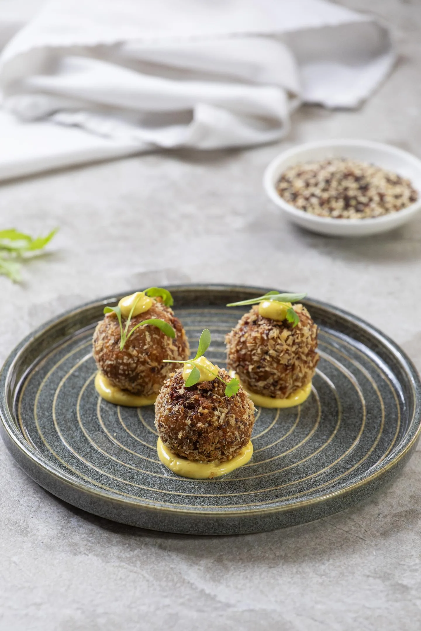 Tres croquetas empanadas, cada una sobre un punto de salsa amarilla y coronada con brotes verdes, en un plato de cerámica gris.
