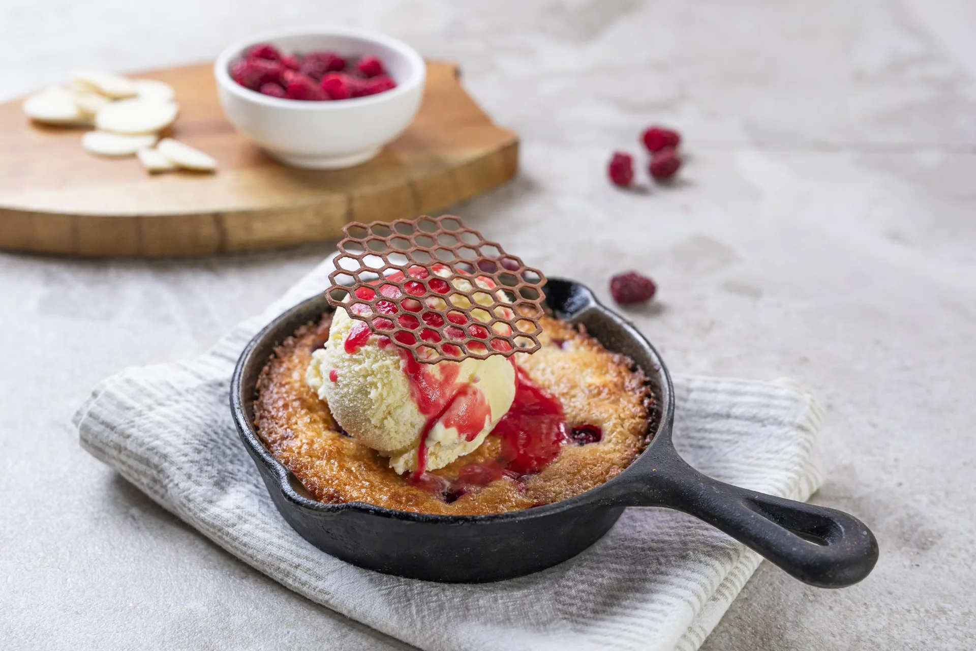 Postre horneado con fruta roja en una sartén de hierro, con una bola de helado y una teja de caramelo encima; al fondo, láminas de almendra y un cuenco de frambuesas.