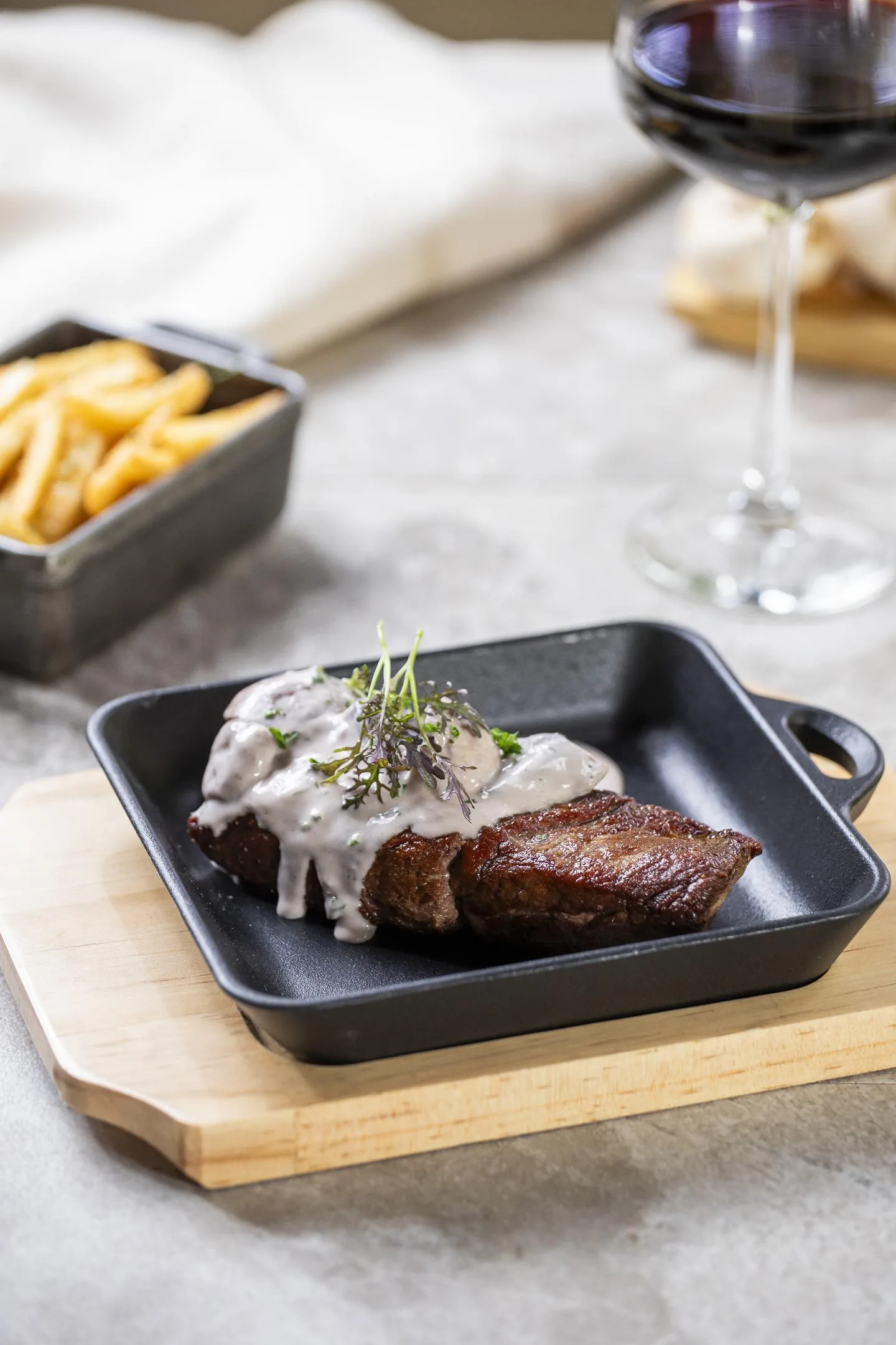 Corte de res cubierto de una salsa cremosa con brotes morados y verdes, en una plancha cuadrada de hierro, con papas fritas y una copa de vino tinto al lado.