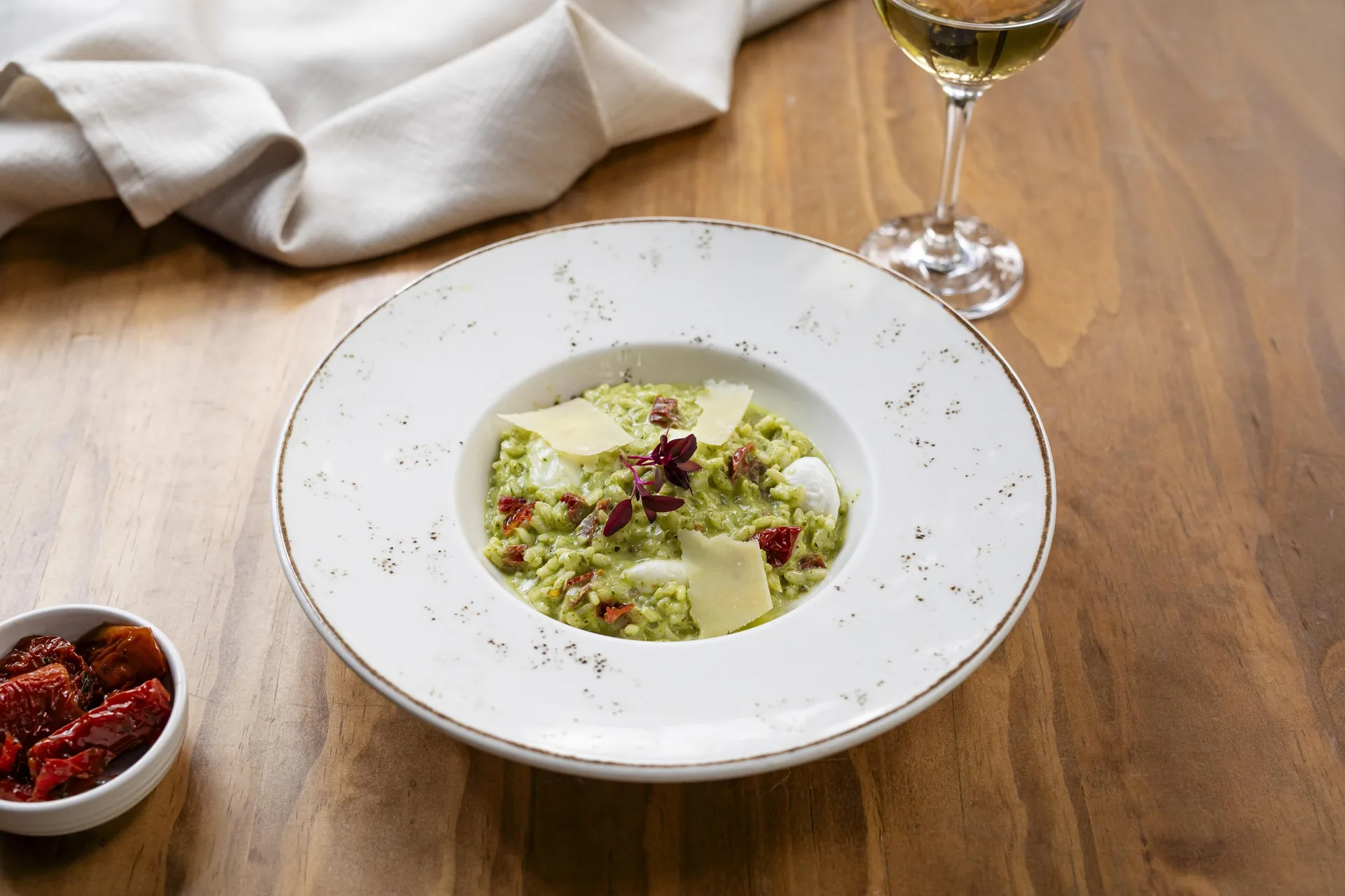 Risotto verde con lascas de queso curado y trozos de tomate seco, en un plato hondo de borde dorado, junto a una copa de vino blanco.