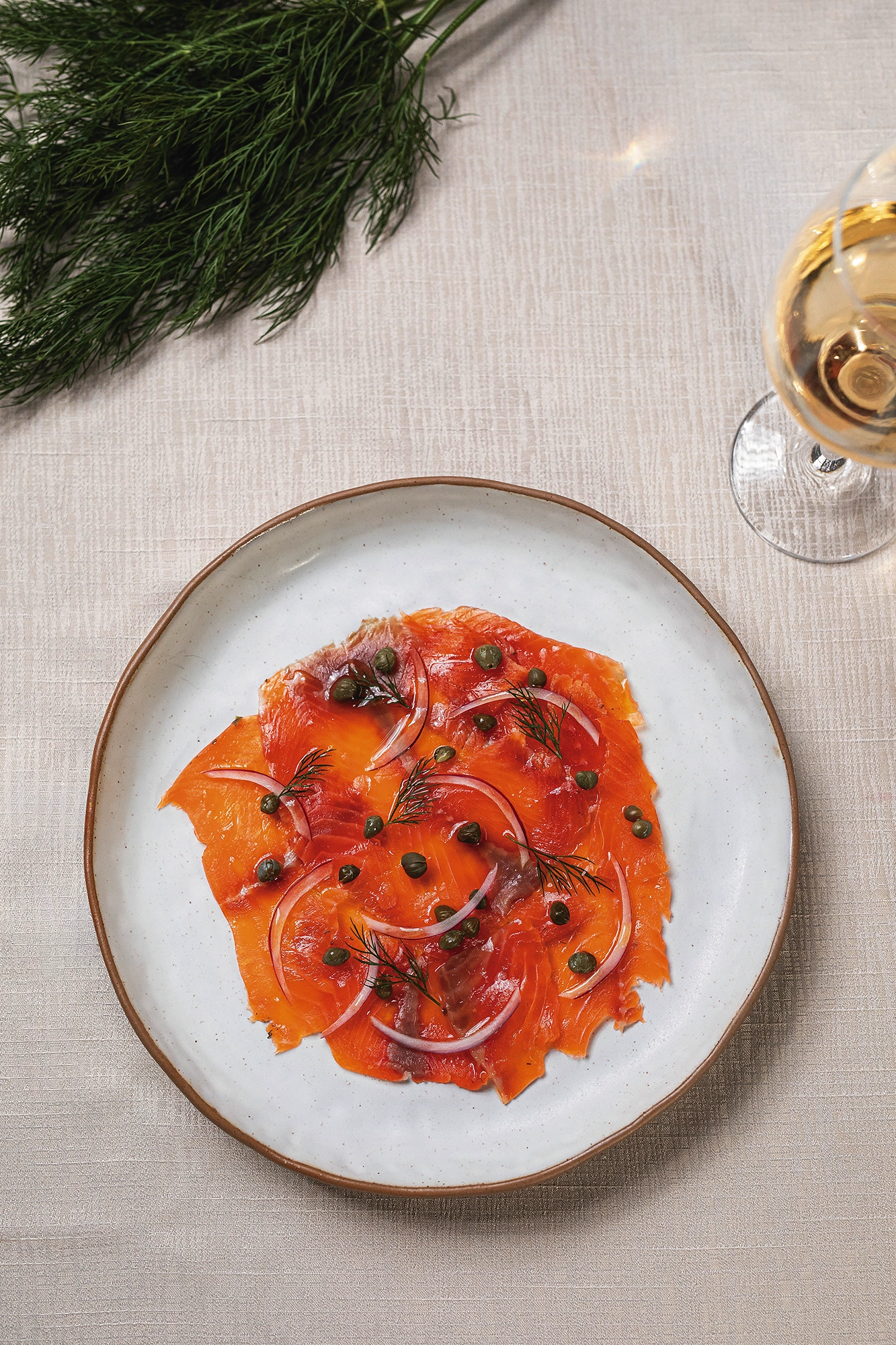 Carpaccio de trucha con alcaparras, aros de cebolla morada y eneldo, en un plato de cerámica junto a una copa de vino blanco