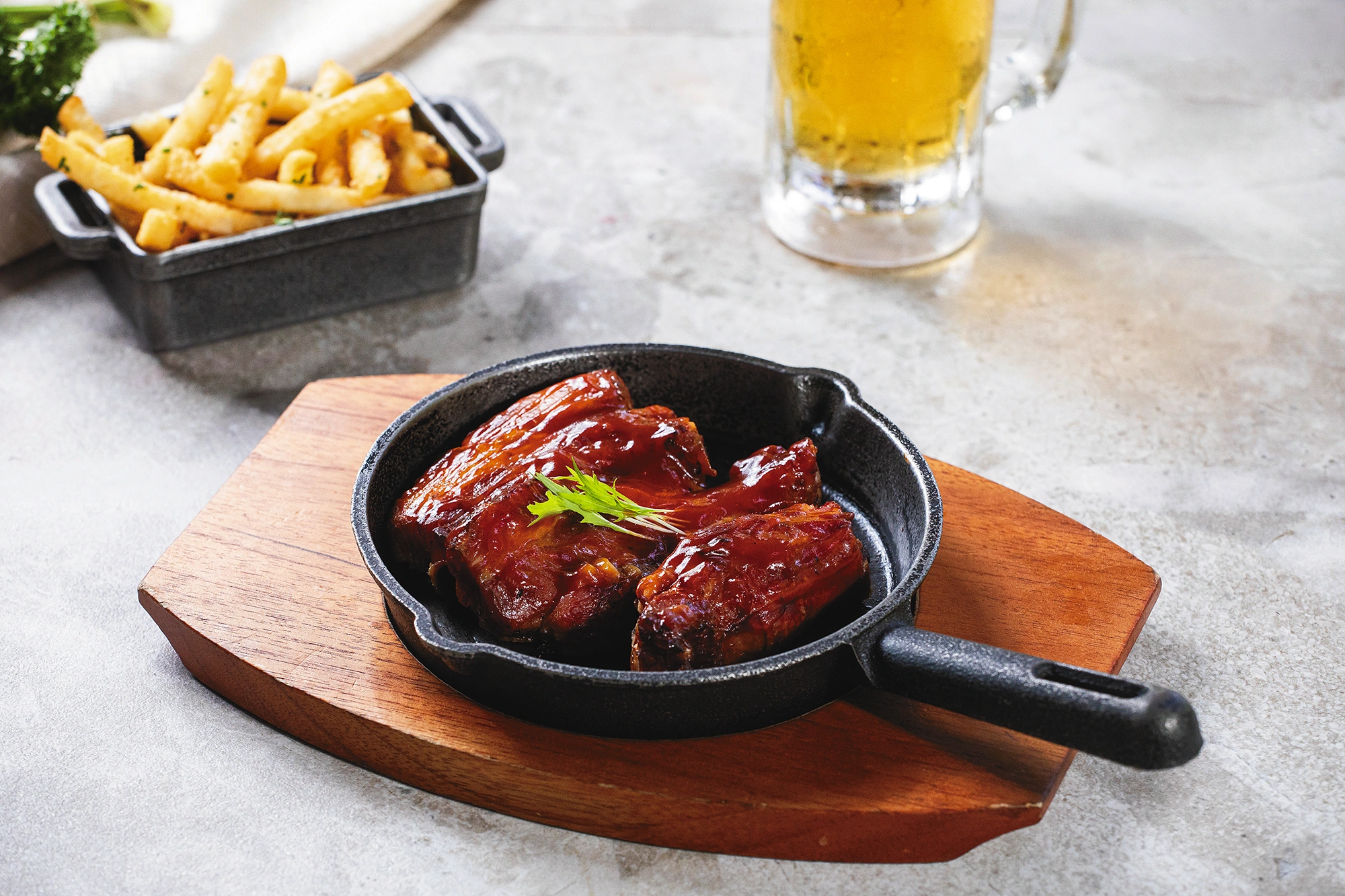 Costillas de cerdo glaseadas en una sartén de hierro sobre tabla de madera, con papas fritas y una cerveza al fondo