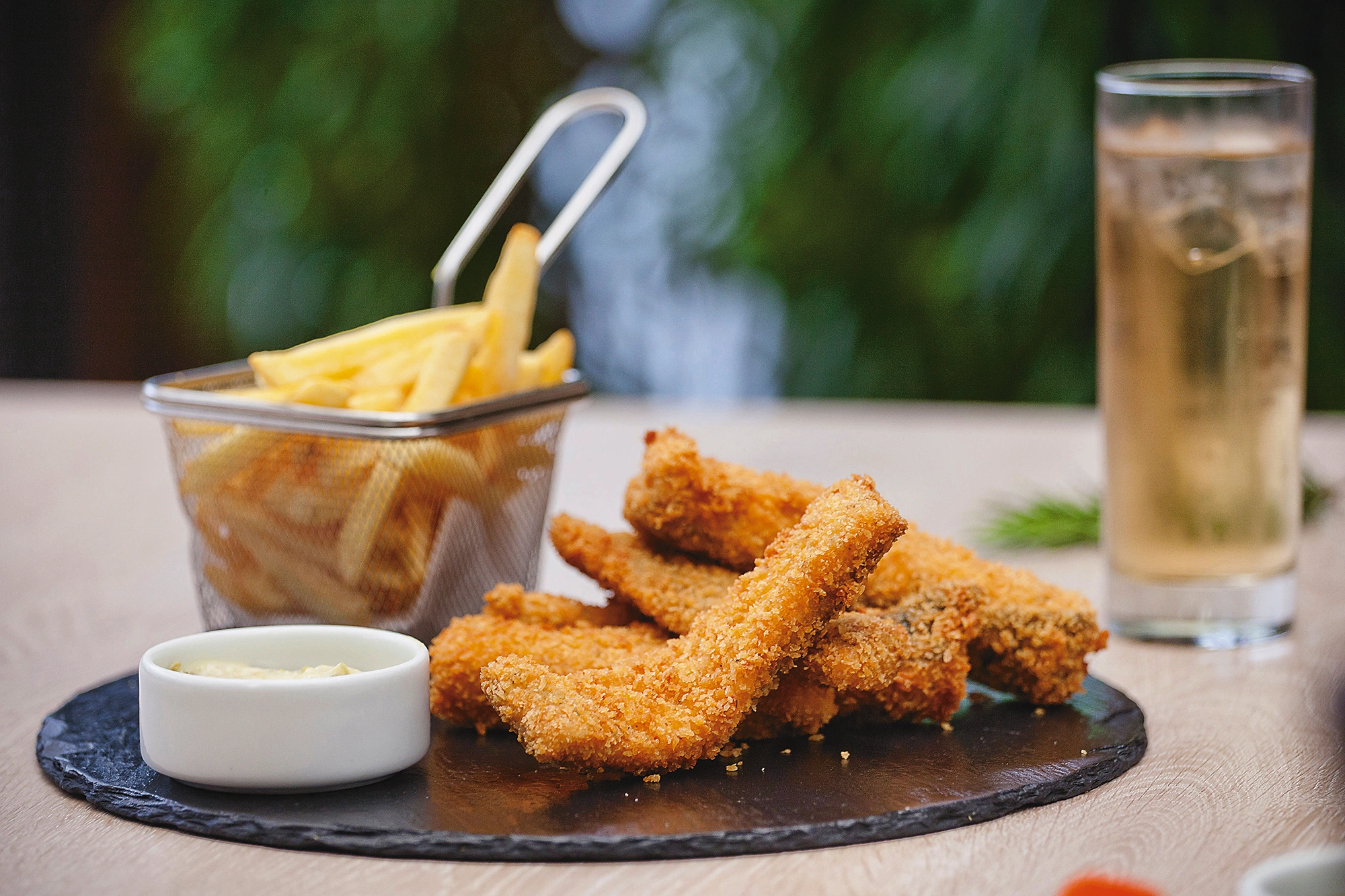Dedos de pollo empanizados sobre pizarra, con papas fritas en cesta metálica y un vaso de bebida fría