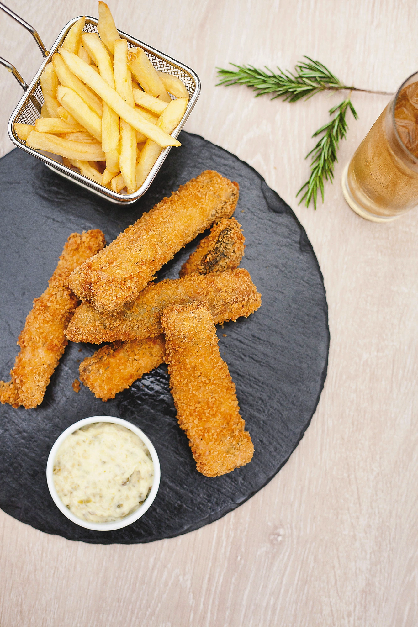 Dedos de trucha empanizados sobre una pizarra redonda, con papas fritas en cesta metálica y salsa tártara