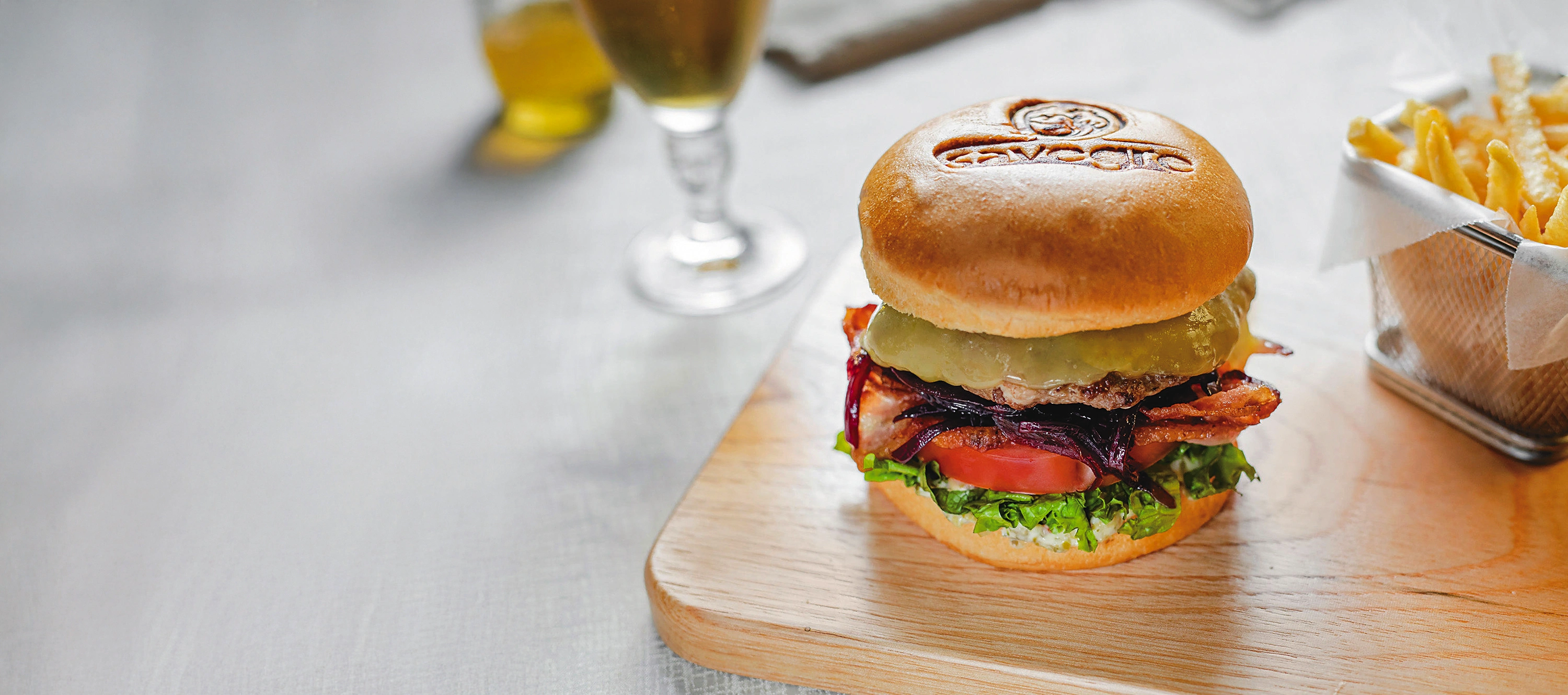 Hamburguesa con el logotipo de Savegre grabado en el pan, sobre tabla de madera, con papas fritas al lado