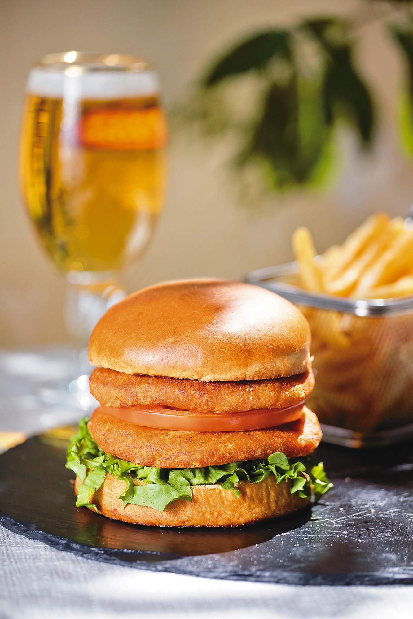 Hamburguesa de trucha con lechuga y tomate en pan brioche, con papas fritas y una cerveza al fondo