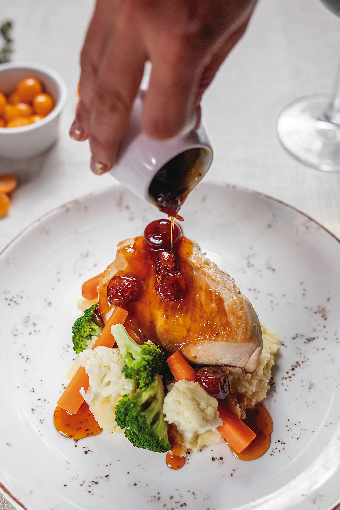 Pechuga de pollo con salsa de frutos rojos que se vierte desde una jarrita, acompañada de puré y verduras
