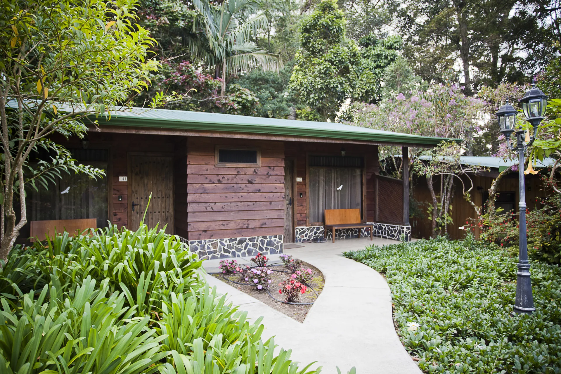 Wooden façade of a room block under a green roof, with a stone-and-wood bench between the two doors and a path running between flower beds.