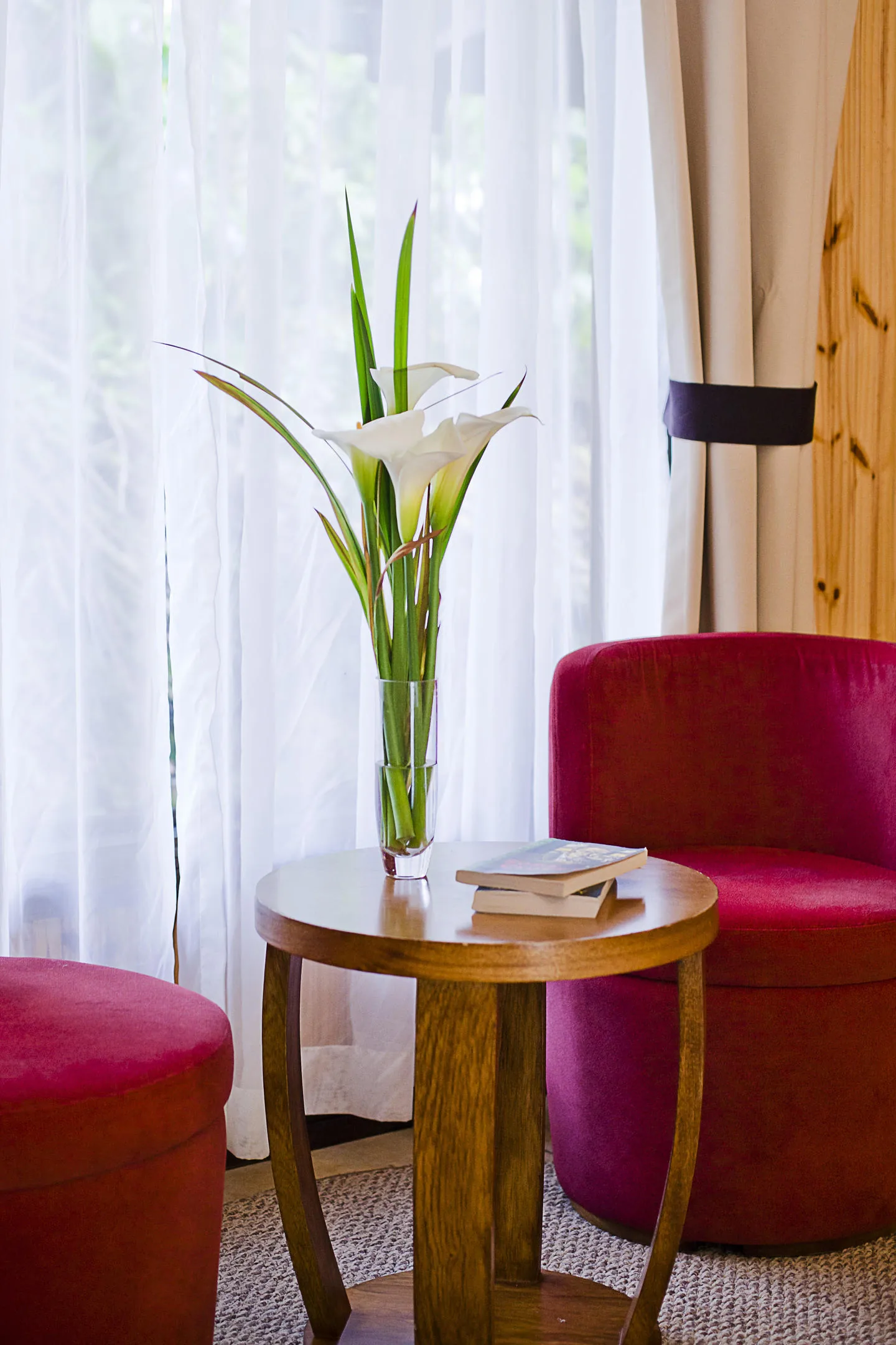 A corner by the window with a round table, three books and a vase of white calla lilies, in front of a raspberry-coloured armchair.
