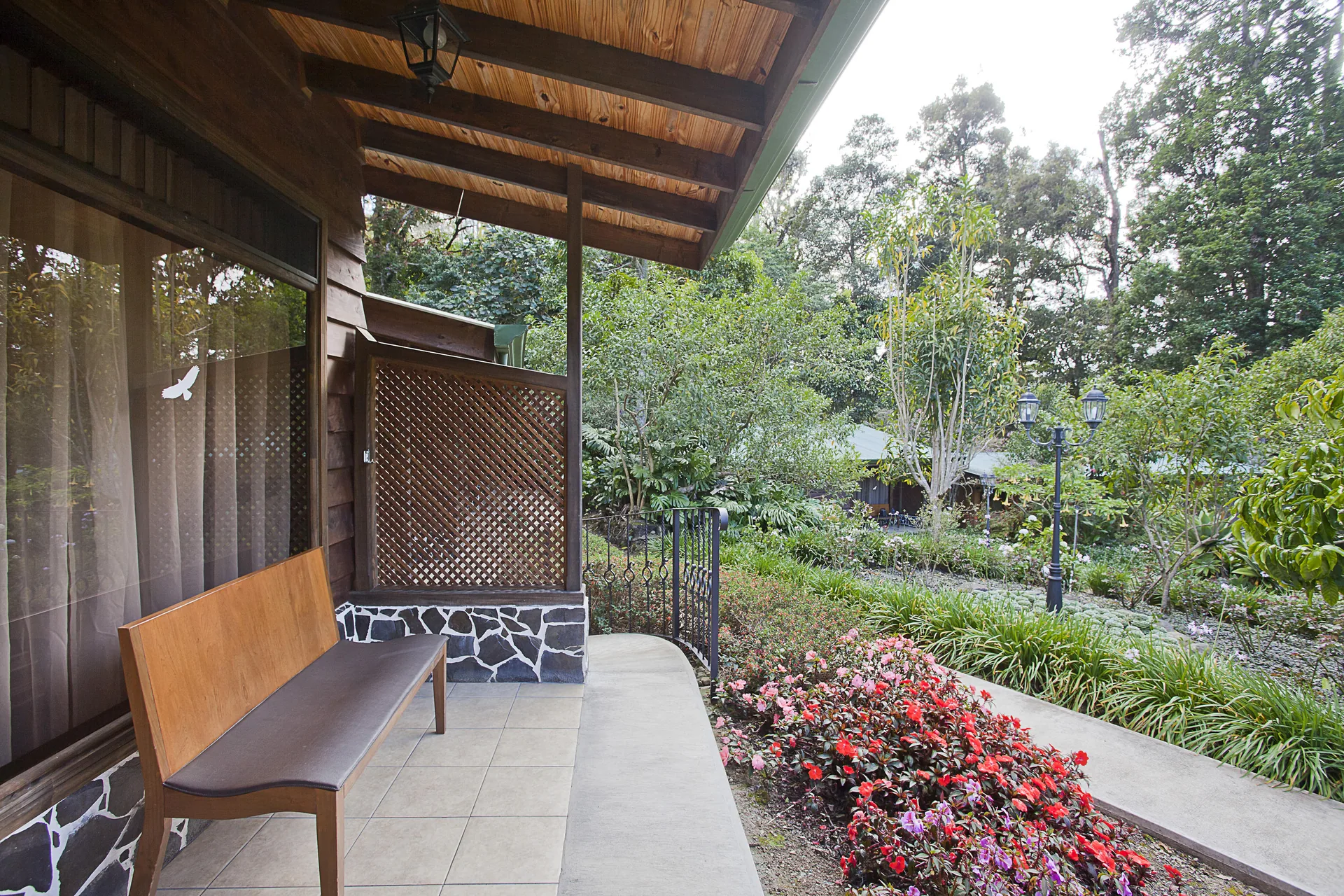 A covered terrace with a beamed wooden roof and a bench against the wall, next to a lattice screen and the flowering garden.