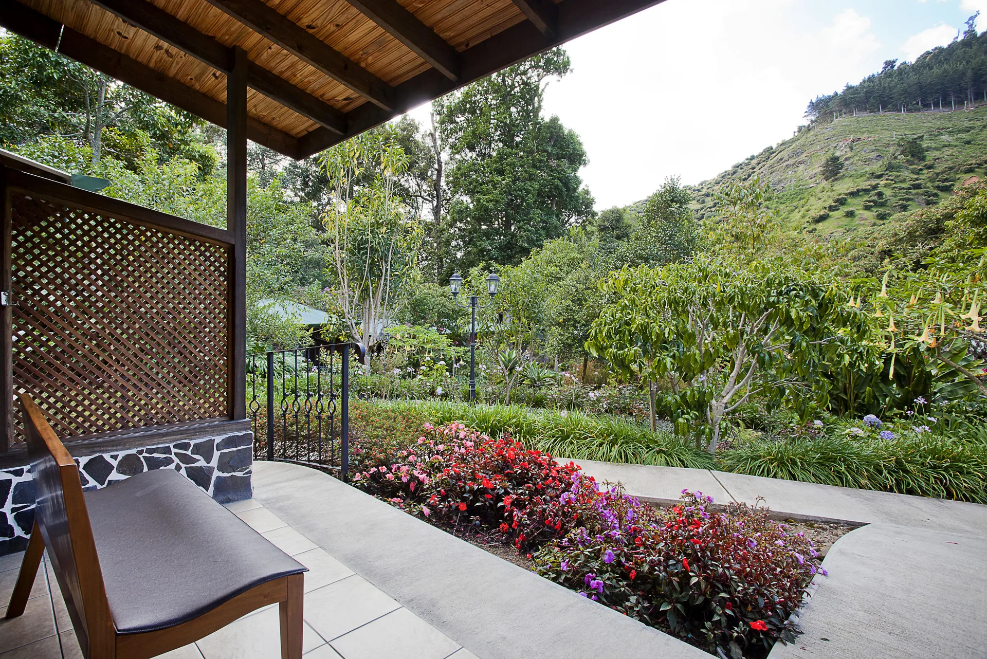 A wooden bench on the terrace at left, a flowering garden filling the middle of the frame and, beyond it, a pasture slope with scattered shrubs and trees along the ridge.