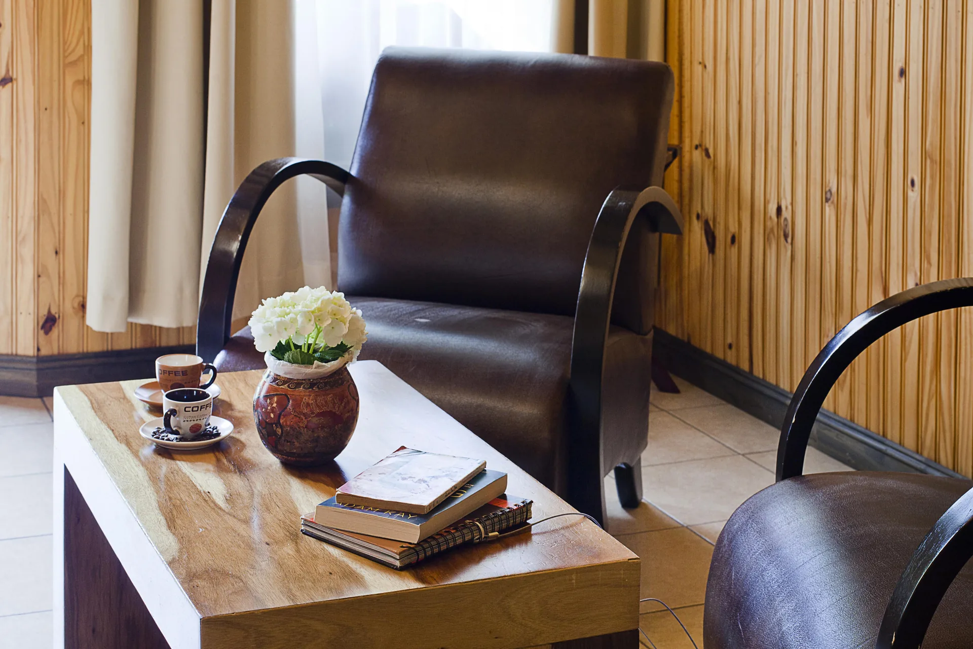 A low table with a vase of white roses, two coffee cups and three books, between two leather armchairs.