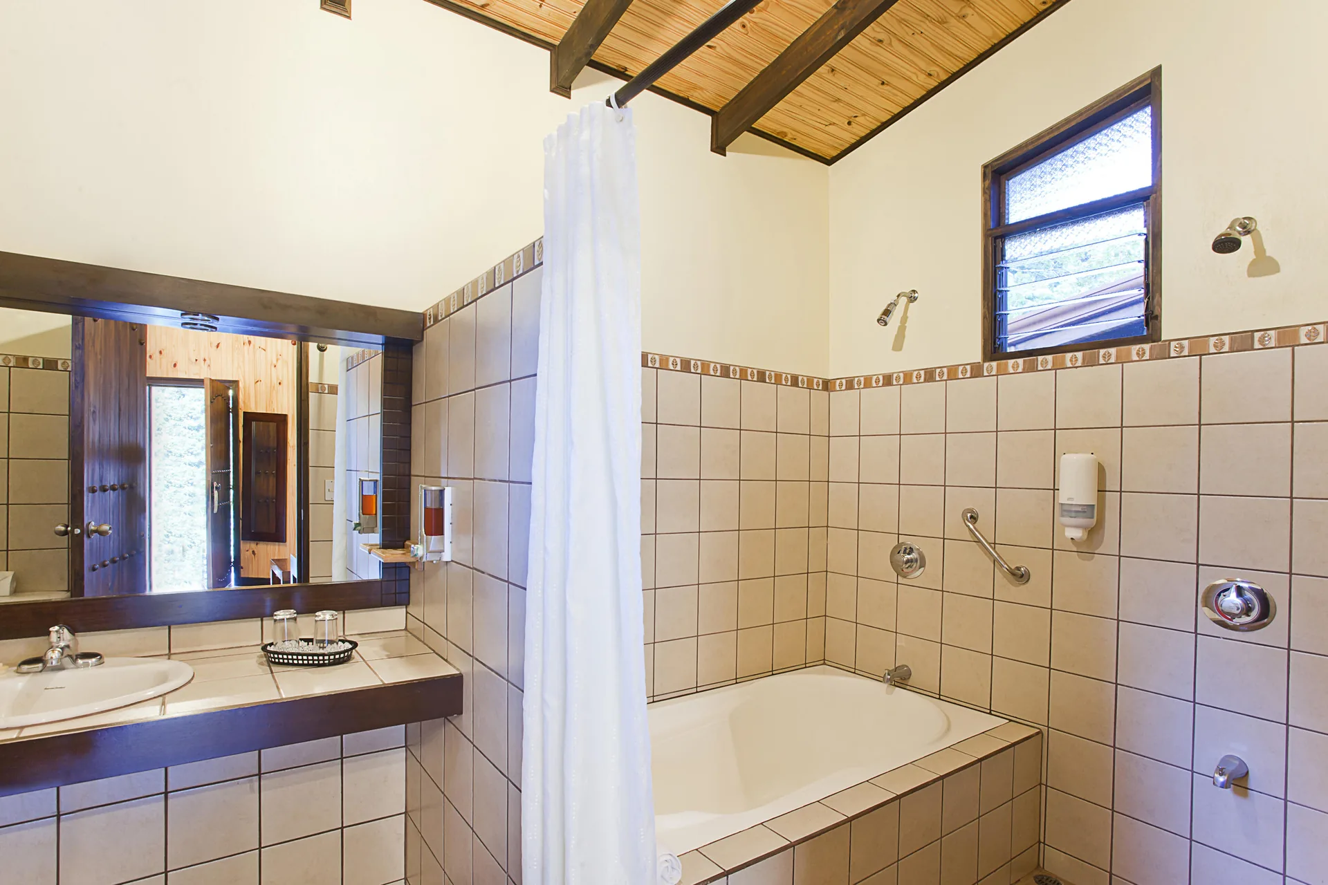 A bathroom with a bathtub and shower behind a white curtain, a washbasin with a mirror and a beamed wooden ceiling.