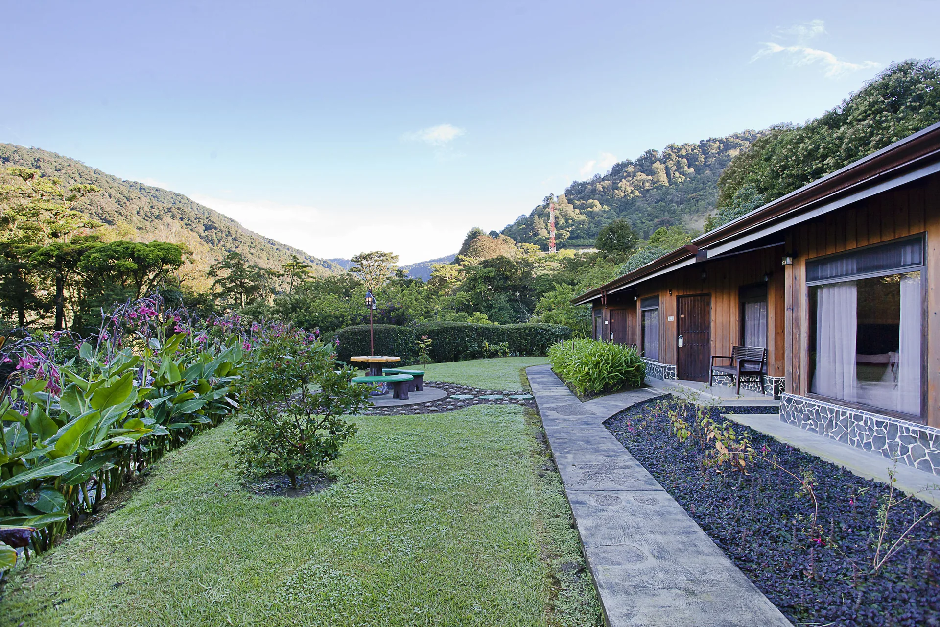 The room block with its long terrace, beside a sloping garden with a picnic table and the forested mountains behind.