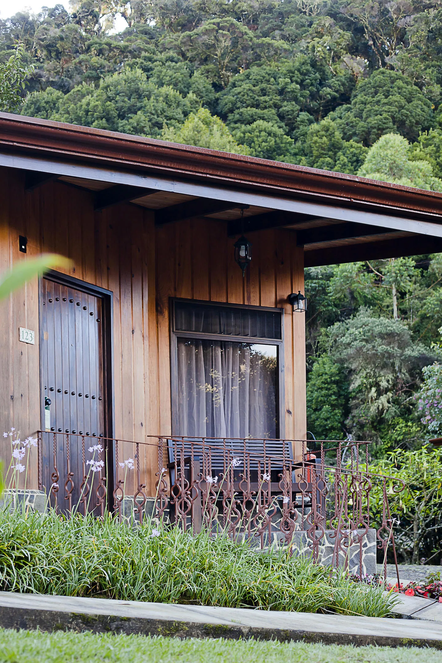The corner of cabin 123, with its terrace bench, a railing and white flowers in front, and dense forest behind.