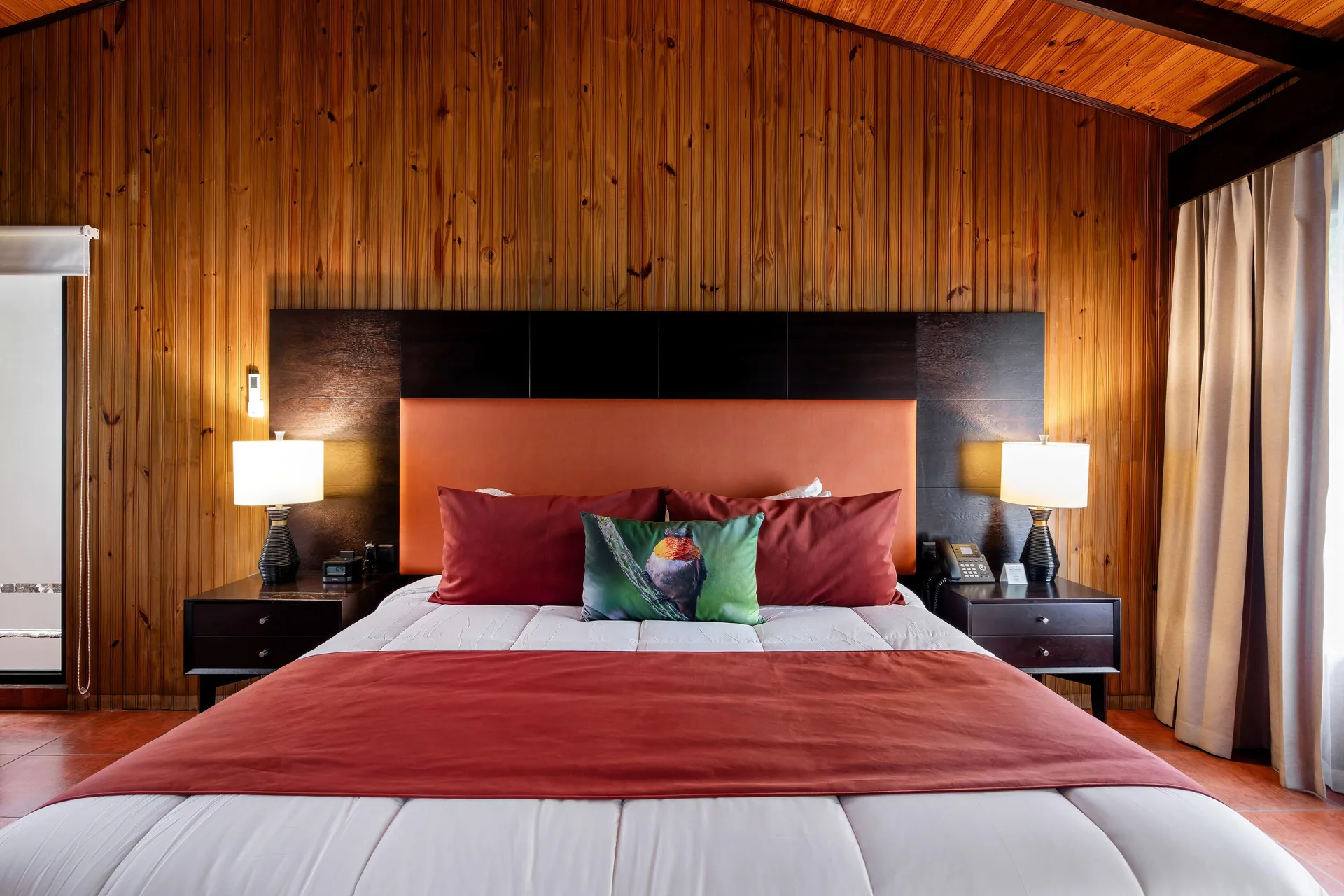 Cama King de la Master Suite con colcha roja, entre dos mesas de noche y bajo las paredes de madera