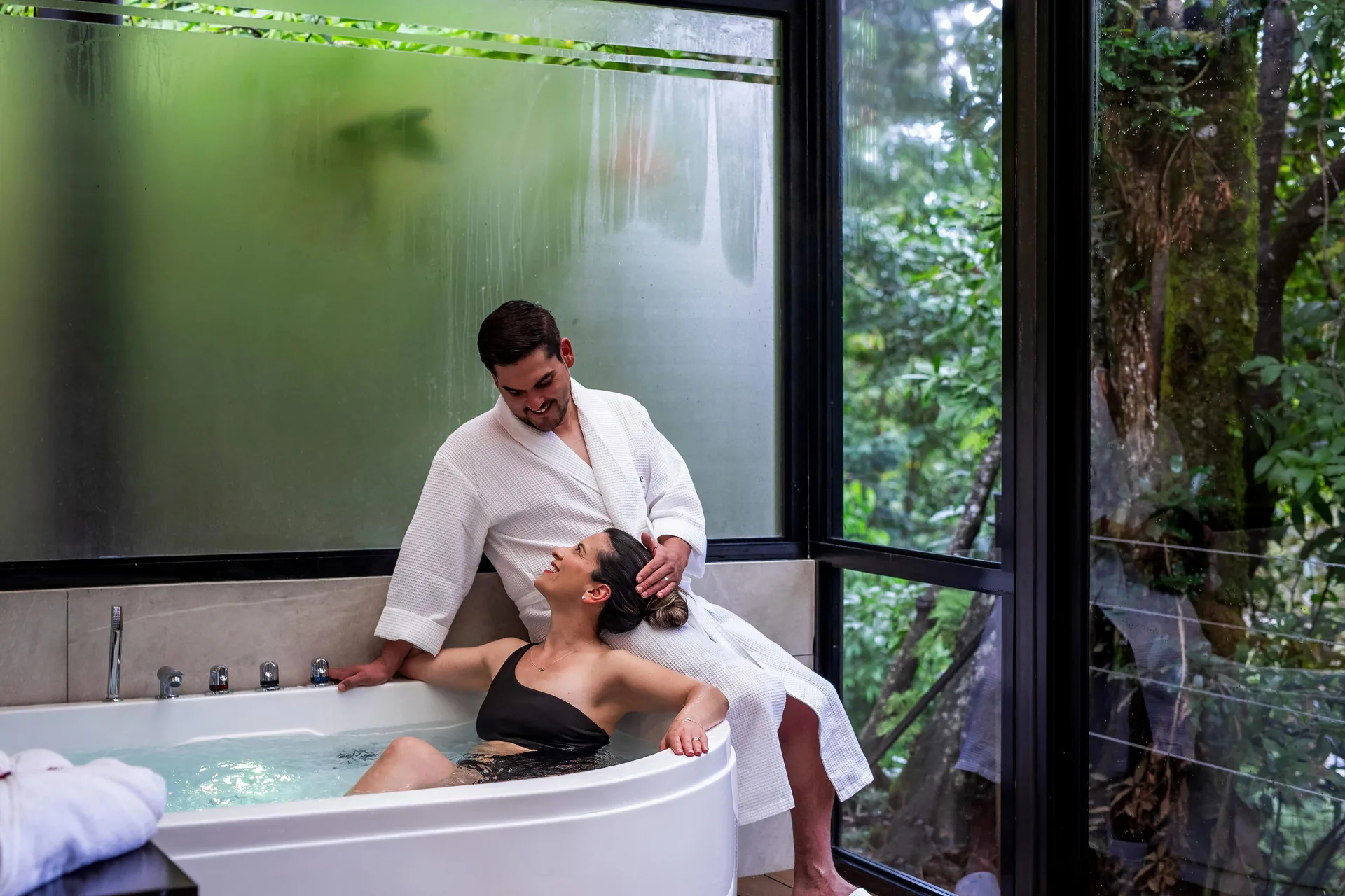 A woman in the jacuzzi and a man in a bathrobe sitting on the edge, beside the window looking onto the forest.