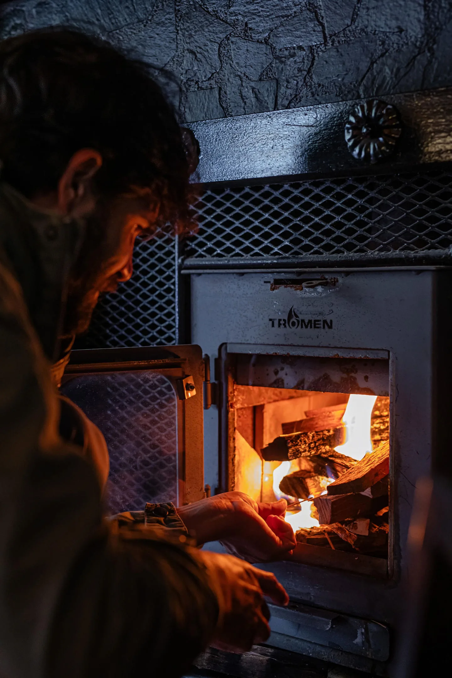 A man lights the wood in the iron stove; the firelight is the only light in the room.