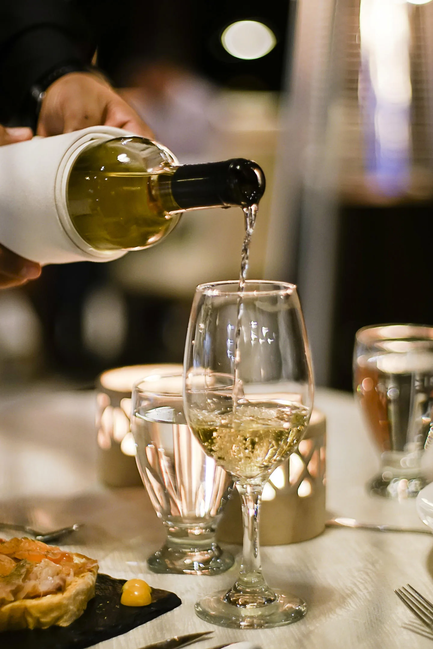 Una mano sirve vino blanco en una copa sobre una mesa vestida, de noche; en primer plano, un aperitivo en un plato de pizarra.