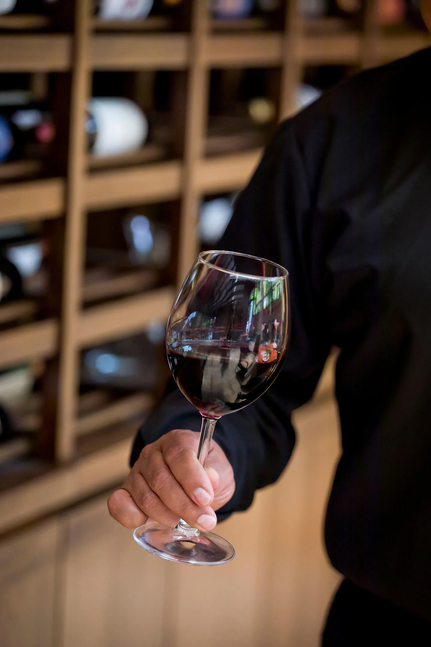 Una mano sostiene por el pie una copa de vino tinto frente al mueble de botellas de la cava.