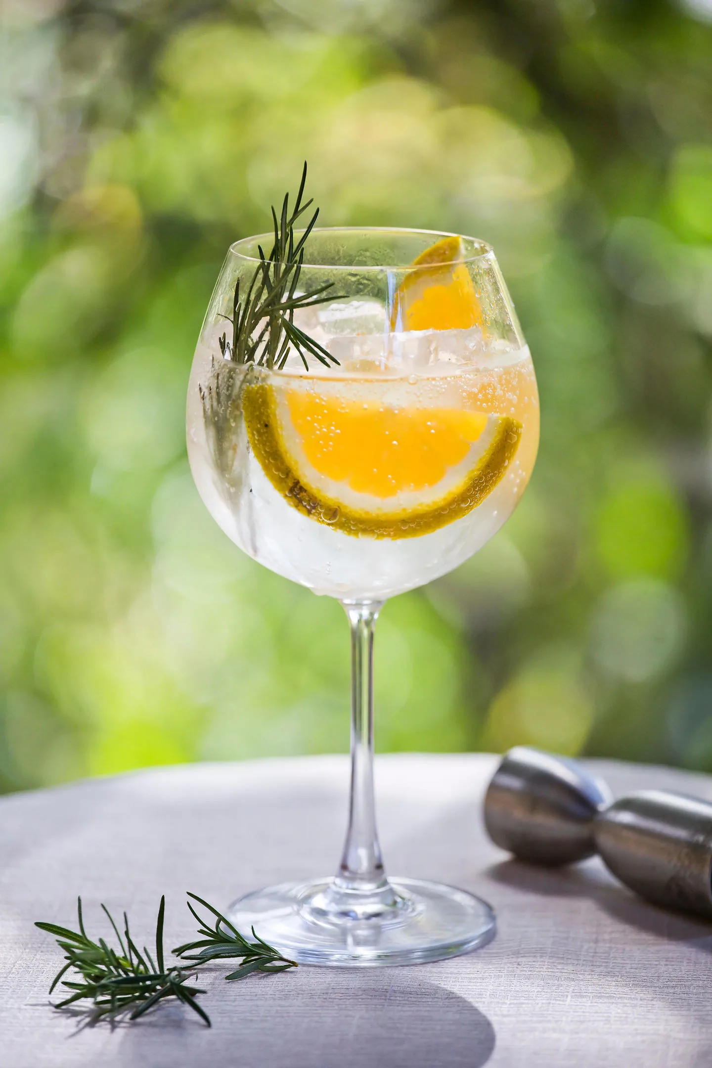 Copa balón con hielo, una rodaja de naranja y una ramita de romero, sobre una mesa clara con la vegetación verde de fondo.