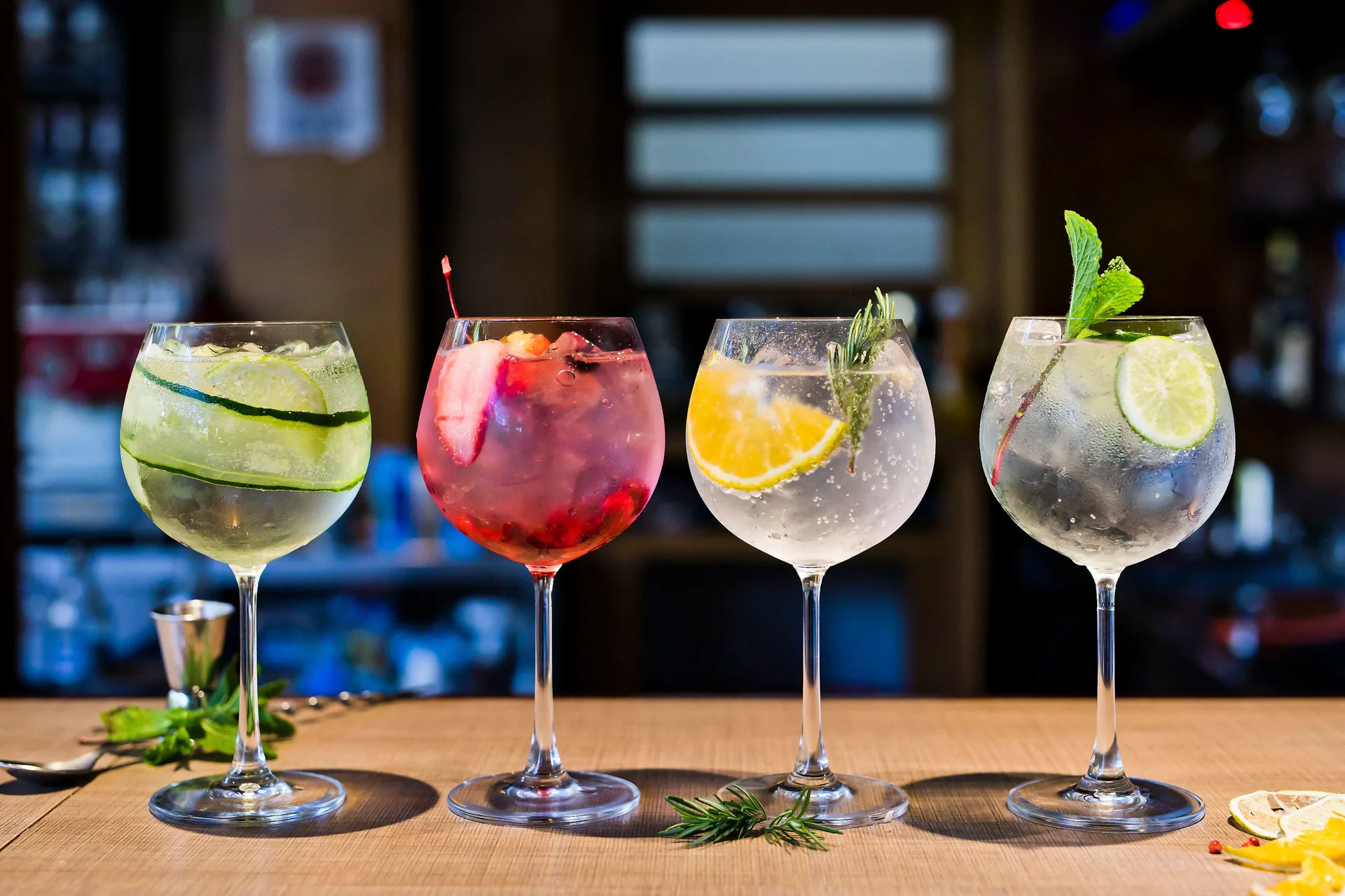 Cuatro copas balón alineadas en la barra, decoradas cada una de forma distinta: pepino en espiral, frutos rojos, naranja con romero, y lima con menta.