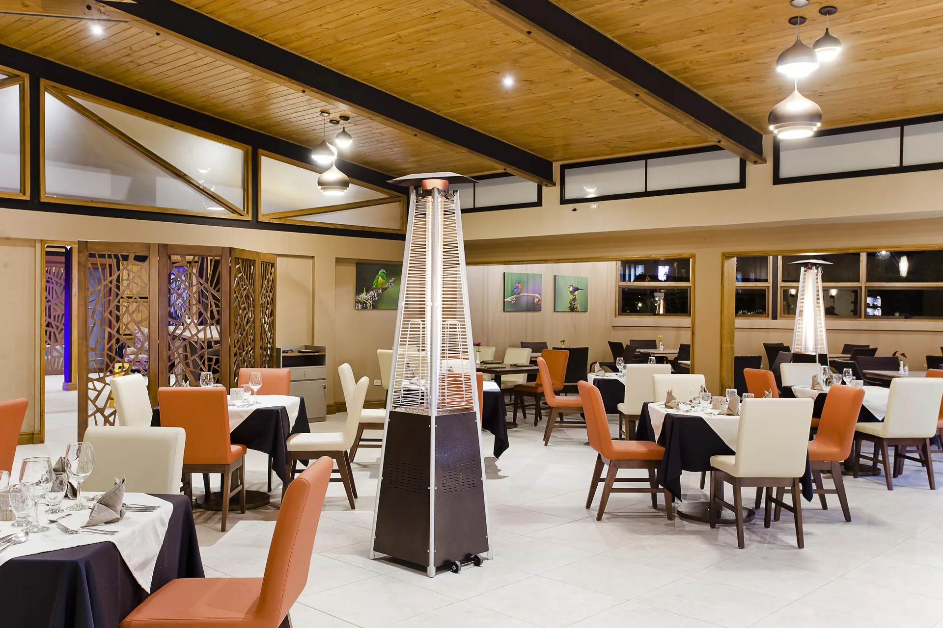 El salón del restaurante, con mesas vestidas de mantel oscuro y sillas naranjas y blancas bajo un techo de madera a dos aguas con vigas negras y lámparas colgantes.