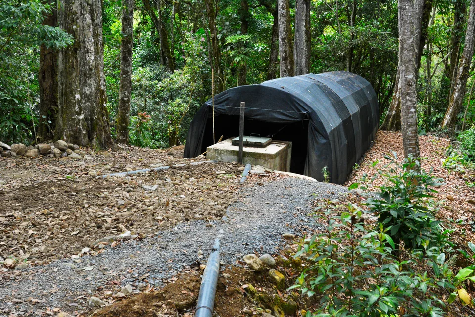 An anaerobic biodigester covered by a dark tarp tunnel, next to a concrete inspection chamber, in the middle of the forest