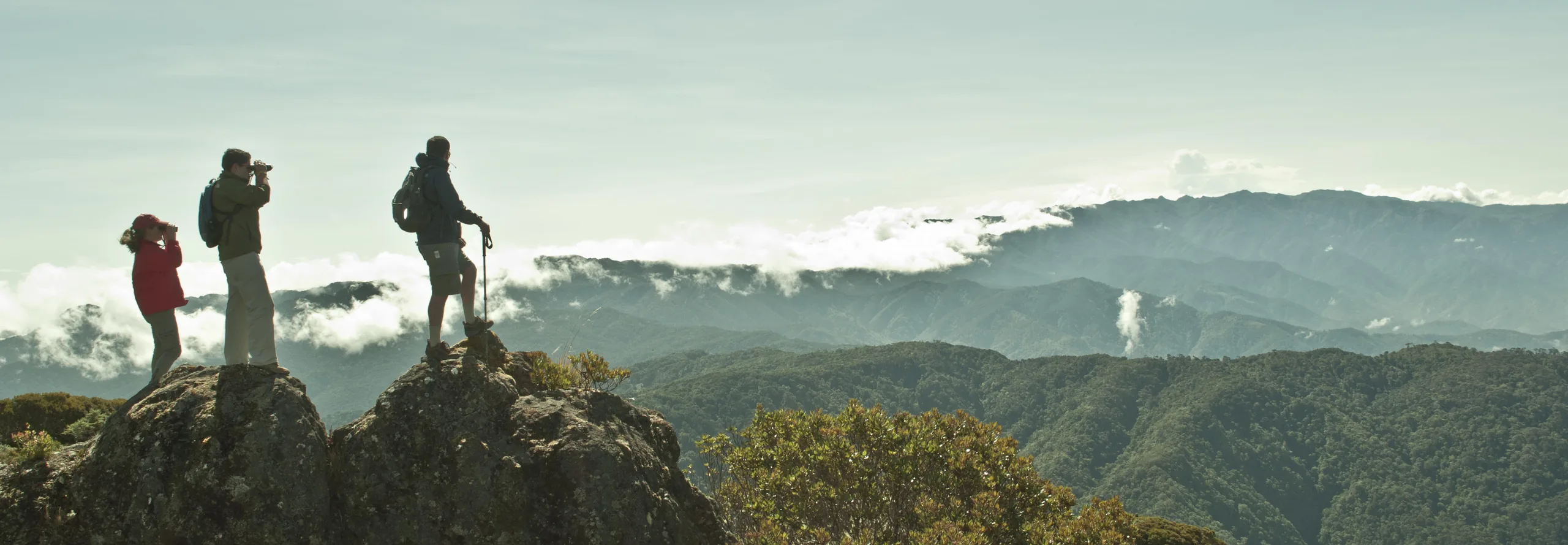 Three figures silhouetted on a rocky outcrop at dawn, one with a camera, above a sea of cloud and ridge after ridge of the mountain range